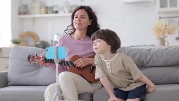 Mãe feliz ligação em casa com o filho pequeno tocar guitarra, cantar músicas e gravar vídeo no smartphone — Vídeo de Stock