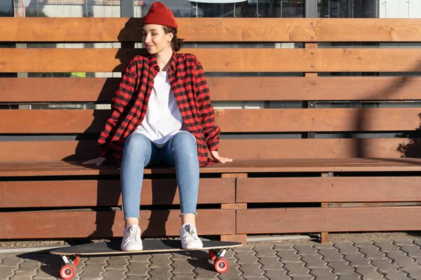 Νέοι hipster κορίτσι skateboarder απολαμβάνουν το φως του ήλιου κάθονται σε ξύλινο πάγκο με τα πόδια σε longboard χαμογελώντας — Φωτογραφία Αρχείου