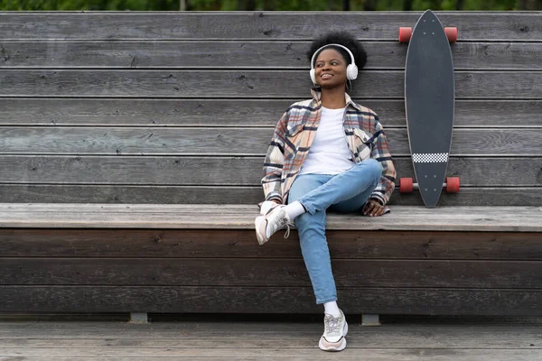 Αφρικανική κοπέλα με longboard ακούστε μουσική με χαρούμενο χαμόγελο μετά από skateboarding chill στο πάρκο εξωτερική — Φωτογραφία Αρχείου