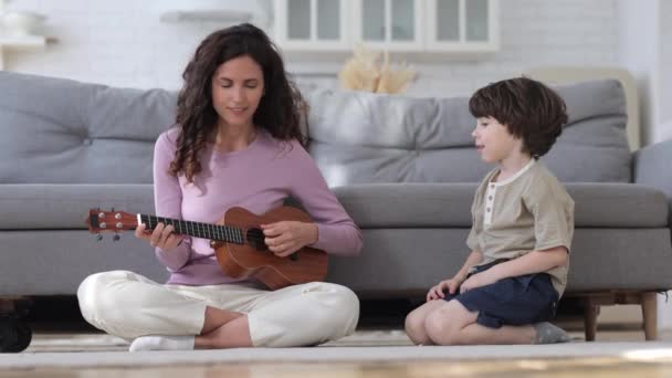 Ciekawy chłopiec, spójrz na mamę trzymającą ukulele. Młoda opiekunka lub nauczycielka uczy dzieci grać na gitarze — Wideo stockowe