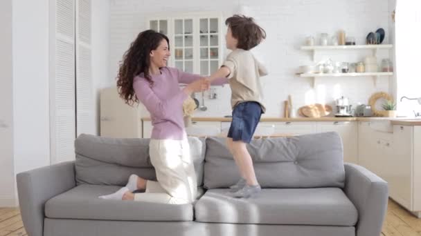 Liten son håller lycklig mamma hand hoppar på soffan. Glada barnvakt eller mor som har roligt med pojken — Stockvideo