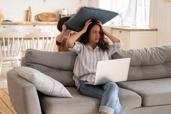 Frustriert mutter versuchen bis arbeit von zuhause sitzen mit laptop auf couch mit kleinem sohn schlagen sie kissen — Stockfoto