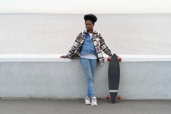 Молода африканська жінка з модним довгим бортом над річкою, що відтворюється після скейтбордингу на набережній — стокове фото