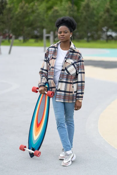 Joven patinadora africana sostiene longboard en skate park. Mujer urbana en ropa casual en skatepark — Foto de Stock