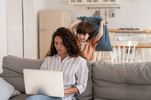 Travail de la maison avec vilain enfant : jeune maman utiliser un ordinateur portable pour les affaires assis sur un canapé avec ennuyé attaque de fils — Photo