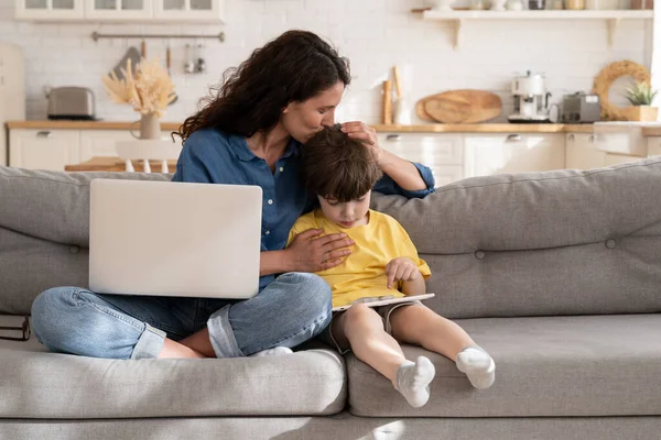 원격 근로자 엄마나 프리랜서는 집에서 일하기 위해 노트북 컴퓨터를 사용하는 태블릿을 가진 아이를 안아 준다 — 스톡 사진