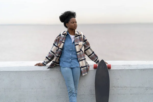 Αφρικανική κοπέλα σταθεί πάνω από το ποτάμι άποψη ματιά στην άκρη κρατήσει longboard φορούν μοντέρνα αστικά ρούχα και αξεσουάρ — Φωτογραφία Αρχείου