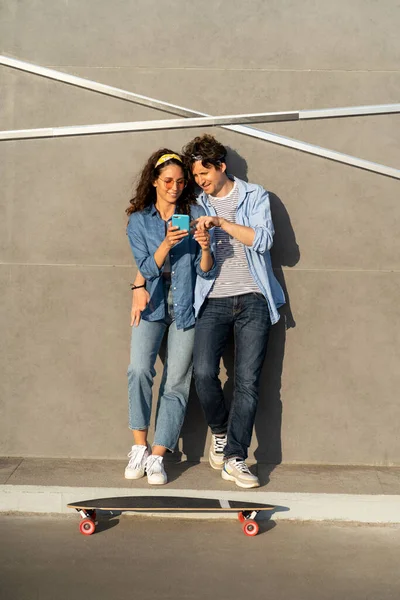 Δροσερό νεαρό ζευγάρι που κοιτάζει στην οθόνη smartphone σε εξωτερικούς χώρους αγκαλιάσει σταθεί σε longboard skate χαρούμενο χαμόγελο — Φωτογραφία Αρχείου