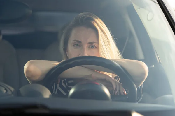 Thoughtful tired woman driving car, looking through windshield at camera, thinking about something.