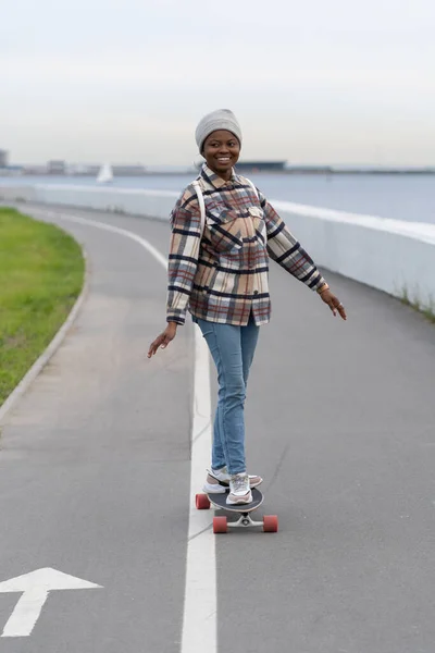 Chica africana sonriente en el monopatín. Happy skateboarder hembra disfrutar de longboarding a principios de primavera. — Foto de Stock