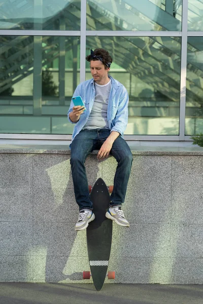 Hipster skater homem usando smartphone segurar longboard na mão ler mensagens no telefone celular online — Fotografia de Stock