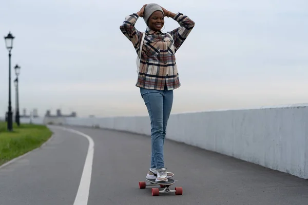 Alegre africano casual chica cabalgando longboard con sonrisa emocionada disfrutar de la actividad de ocio al aire libre — Foto de Stock