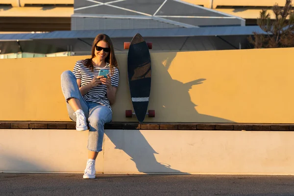 Joven europeo chica chat usando smartphone aplicación sentado con longboard skate en parque al atardecer — Foto de Stock