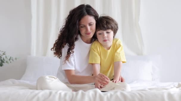 思いやりのあるお母さん座って上のベッドとともに就学前の子供の本を読むか、画像を表示するには小さな男の子説明教育 — ストック動画
