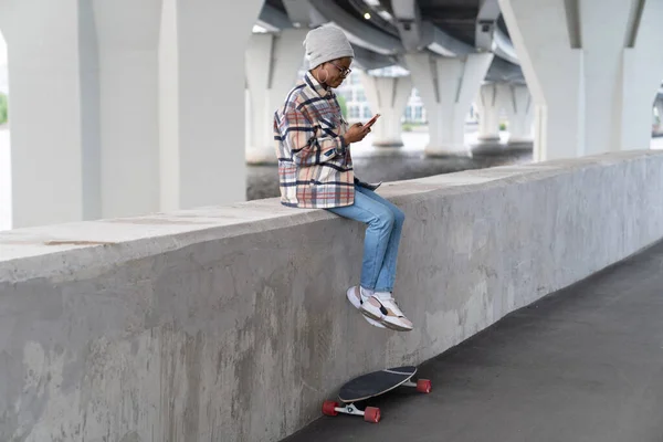 Casual african skateboarder girl with smartphone in hands sitting on concrete pavement read message — Stok fotoğraf