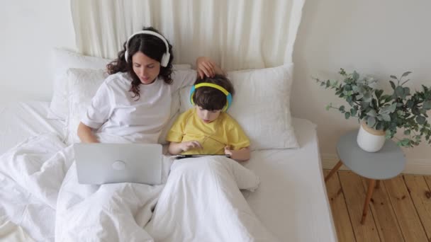 Mãe feliz gosta de trabalhar a partir de casa laptop uso deitado na cama com o pequeno filho assistir desenhos animados em tablet — Vídeo de Stock