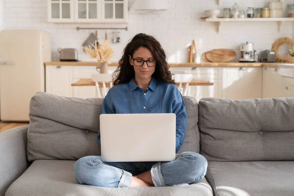 Brunette vrouw in bril gebruik laptop computer zitten op de bank in de woonkamer werk op afstand van huis — Stockfoto