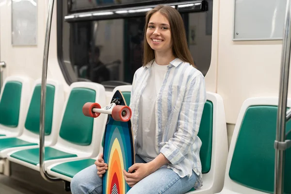 Міленіалка тримає довгу дошку, сидячи в поїзді метро, посміхаючись, дивлячись на камеру. Міське хобі . — стокове фото