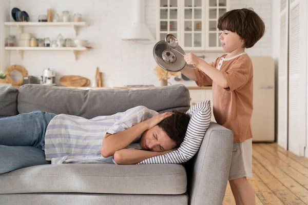 Cansado agotado madre cubre las orejas acostado en el sofá con aburrido hijo ruidoso hacer ruido ruidos utensilios de cocina —  Fotos de Stock