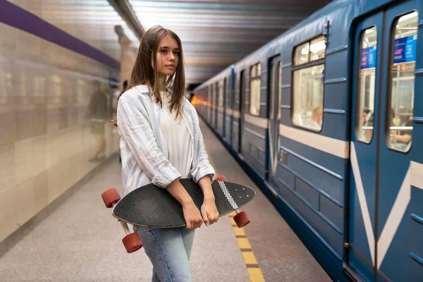 Chica joven en la plataforma del metro mira el coche del metro en la estación de metro subterráneo solo sosteniendo longboard — Foto de Stock