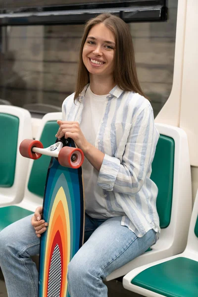 Femme millénaire tenant longboard assis dans le métro train, souriant, regardant la caméra. Loisirs urbains. — Photo