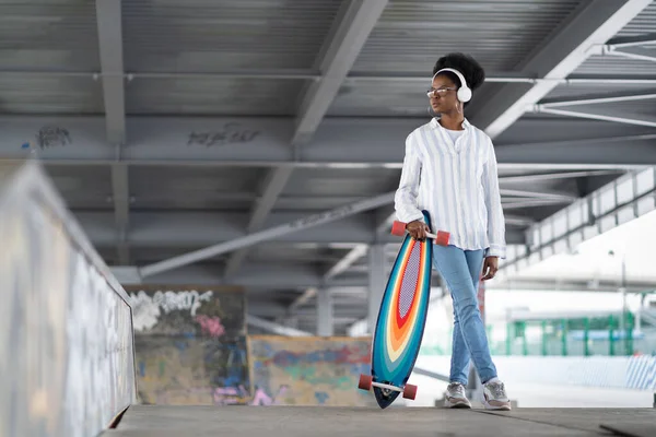 Μοντέρνο casual μαύρη γυναίκα στα ακουστικά κρατήσει longboard στο skatepark ή μοντέρνο αστικό χώρο φόντο — Φωτογραφία Αρχείου