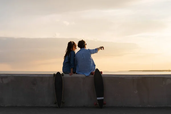 Κομψό ζευγάρι, skateboarders άνδρας και γυναίκα χαλαρώνουν απολαμβάνοντας το ηλιοβασίλεμα κάθονται στο πεζοδρόμιο του παραλιακού δρόμου — Φωτογραφία Αρχείου