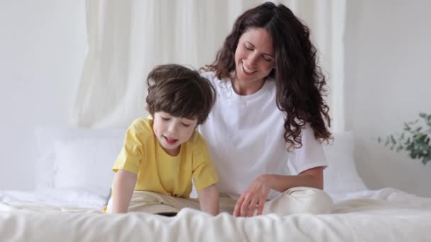 Giovane mamma genitore aiutare il figlio a imparare a leggere la mattina del fine settimana. Mamma sedersi con libro e bambino sul letto — Video Stock