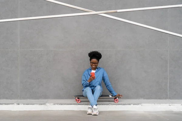 Щаслива чорна дівчина набирає повідомлення в смартфоні або прокручує соціальні мережі через 5g Інтернет, сидячи на відкритому повітрі — стокове фото