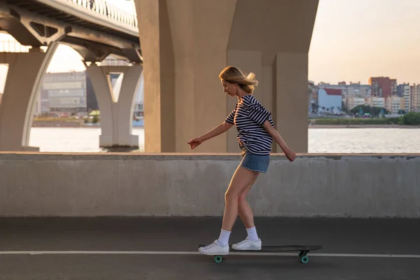 Mujer activa patinando. Mujer rubia en ropa de estilo callejero montando longboard al atardecer en camino vacío — Foto de Stock