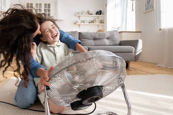Joyful single mother bonding with son excited together at home with fan blowing wind in living room — Stock Photo, Image