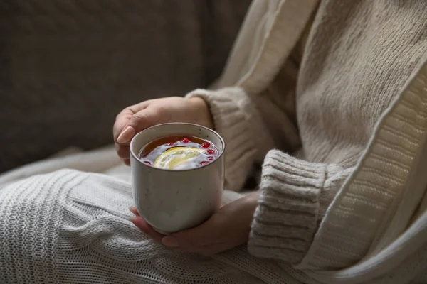 Winter season concept. Woman in wool sweater, covered with a warm blanket,   holding a cup of hot tea with slice of lemon and cranberries. Natural remedies for colds. Cozy Tea Party indoor