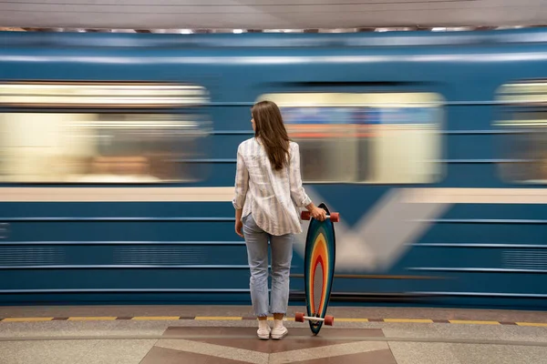 Chica con longboard de pie en la plataforma subterránea con borroso tren azul en movimiento en el fondo — Foto de Stock