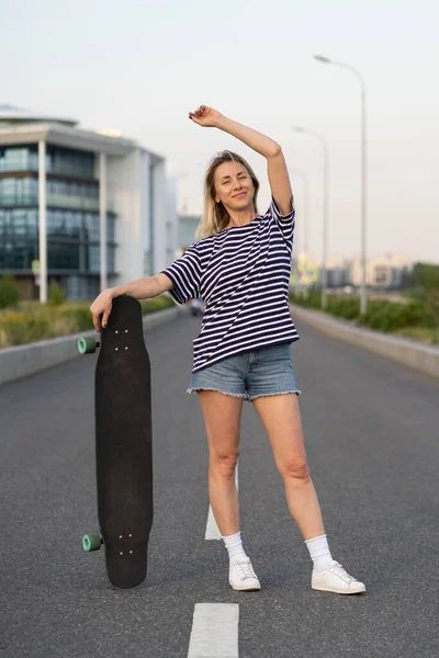 Ενήλικες γυναίκα απολαμβάνουν πατινάζ σε longboard σε άδειο δρόμο της πόλης. Χαρούμενη χαρούμενη γυναίκα κρατώντας skateboard — Φωτογραφία Αρχείου