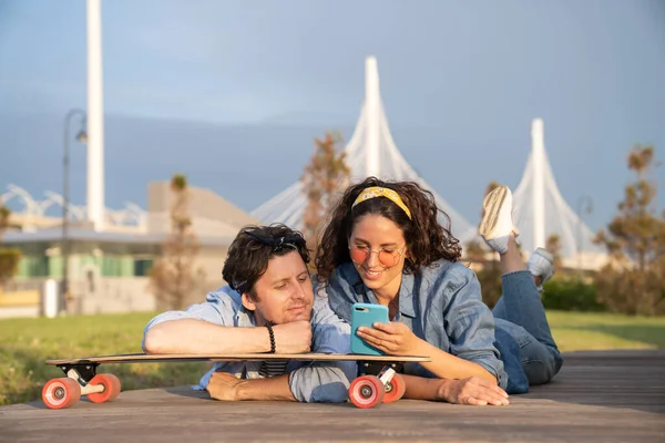 Trendy couple browsing web on smartphone outdoors using 4g internet connection lying on longboard