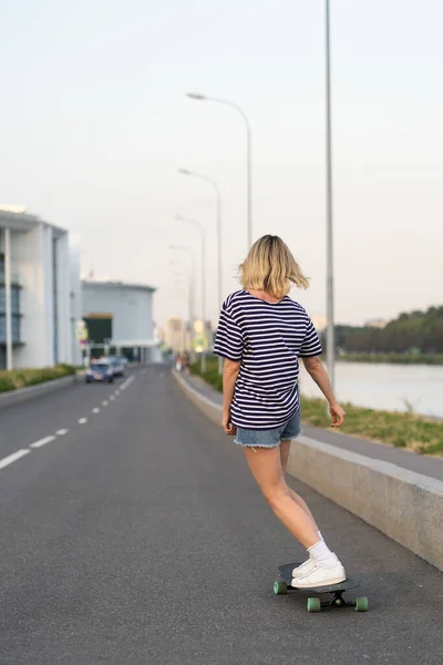 Chica patinadora patinando en la carretera. Mujer rubia en camiseta y pantalones vaqueros cortos en longboard, vista trasera — Foto de Stock