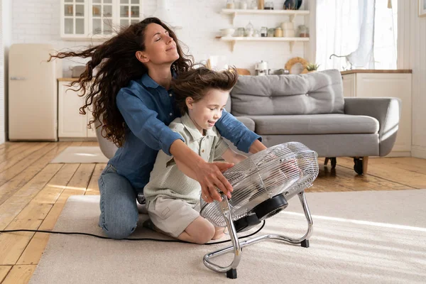 Carefree young mom or nanny have fun with boy son at home sitting together at big fan blowing air — Stock Photo, Image