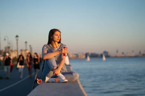 Joven estudiante con monopatín y bebida relajarse mirando la puesta de sol en la orilla del río y disfrutar del verano — Foto de Stock