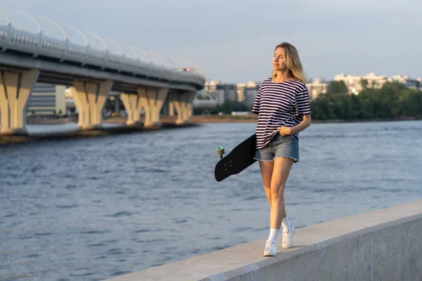 Mujer caminar por el terraplén del río mantenga longboard disfrutar de la puesta de sol de verano en la ciudad. Estilo de vida urbano — Foto de Stock