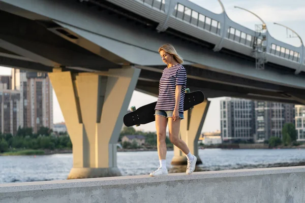 Estilo de vida de la ciudad: paseo de skate femenino a lo largo del río al atardecer sobre el paisaje urbano con puente — Foto de Stock