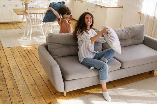 Joven madre risa jugar con el hijo preescolar en casa. Mamá o niñera disfrutan de la pelea de almohadas con el niño — Foto de Stock