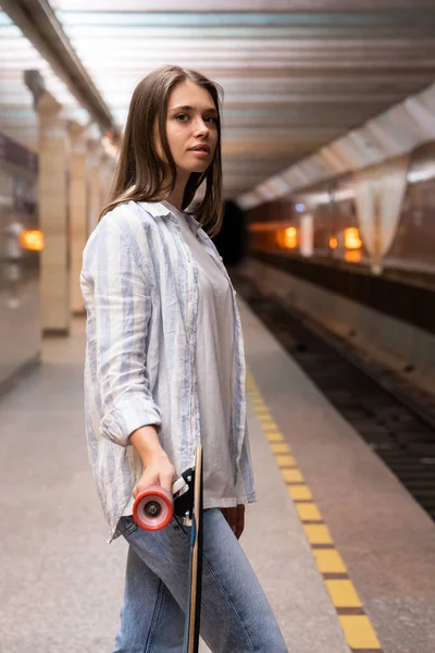 Chica joven con longboard esperar tren en la estación de metro. Mujer en plataforma subterránea con patín — Foto de Stock