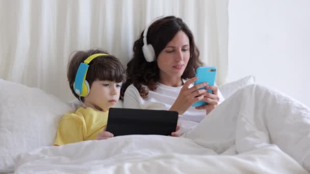 小男孩抱着平板电脑躺在床上，带着耳机和妈妈一起用智能手机听音乐 — 图库视频影像