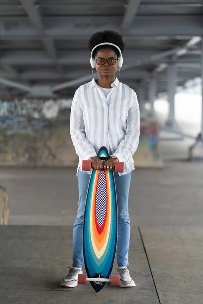 Fille africaine avec skateboard. Femme à la mode habillée en tenue urbaine porter des écouteurs, tenir longboard — Photo