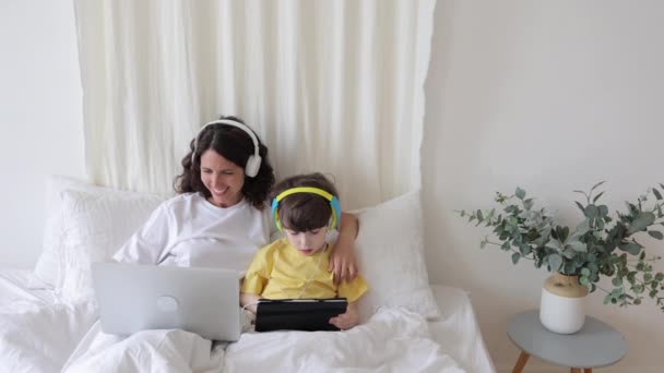 Feliz mañana de los padres con el niño en el dormitorio: mamá joven usa el ordenador portátil abrazando al niño pequeño con la tableta — Vídeo de stock