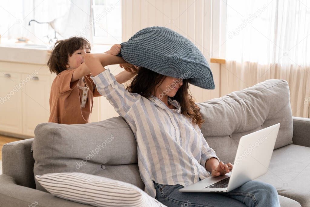 Frustrated mom try to work from home sit with laptop on couch with little son hitting her pillow