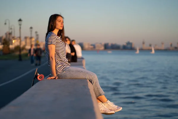 Chica patinadora adolescente en la puesta de sol junto al mar. Mujer joven disfrutar de la noche de verano en el río o el mar mirar el agua — Foto de Stock