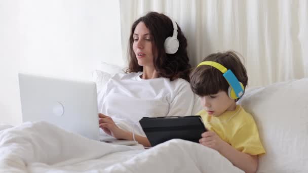Radostná matka dělat nákupy na notebook ležící v posteli s malým synem hodinky karikatury na digitálním tabletu — Stock video