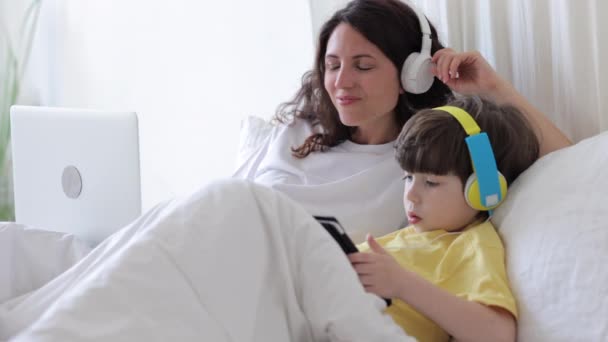 Praca zdalna i edukacja: mama i syn leżący w łóżku z laptopem i robiący lekcje online w tablecie — Wideo stockowe