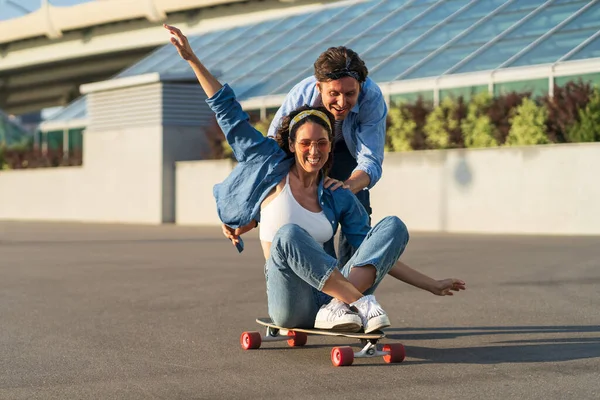 Пара розважається на дошці: чоловіки відштовхуються від радісної жінки, сидячи на скейтборді і сміючись — стокове фото